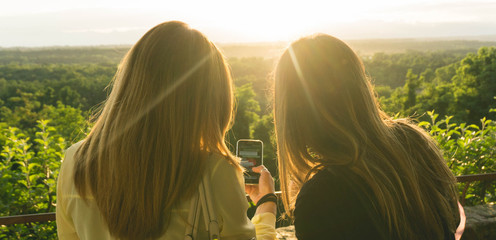 Rear view of couple female friends looking smartphone at sunset. Girlfriend watch cell phone whit...