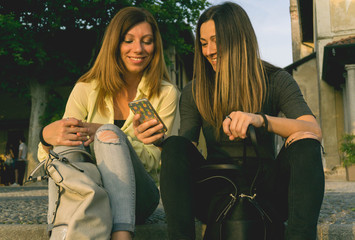 Two beautiful smiling woman look at smartphone in a city park sitting at sunset moment. Woman and the other brunette flip through the photos on the cell phone.