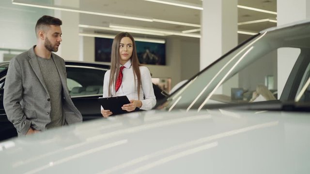 Attractive female car dealer is talking to interested customer about new automobile model and holding documents while standing together near beautiful car.