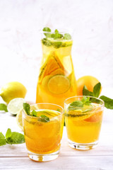Two glasses & pitcher bottle of homemade refreshing lemonade with slices of organic ripe lemon, orange and lime, mint & ice on rustic white wooden table background. Close up, top view, copy space.