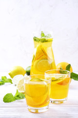 Two glasses & pitcher bottle of homemade refreshing lemonade with slices of organic ripe lemon, orange and lime, mint & ice on rustic white wooden table background. Close up, top view, copy space.