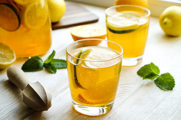 Iced lemonade pitcher & two glasses, wooden juicer, cold citrus infused water, lemon & lime, mint leaves, cutting board on white wooden windowsill. Apartment window background, close up, copy space.