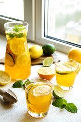 Iced lemonade pitcher & two glasses, wooden juicer, cold citrus infused water, lemon & lime, mint leaves, cutting board on white wooden windowsill. Apartment window background, close up, copy space.