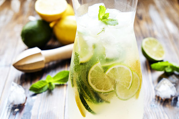 Iced lemonade pitcher, wooden juicer, cold citrus infused water beverage with lemon & lime slices, mint leaves on brown grunged wood textured table sunny day, grunged background. Close up, copy space.