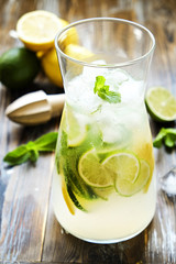 Iced lemonade pitcher, wooden juicer, cold citrus infused water beverage with lemon & lime slices, mint leaves on brown grunged wood textured table sunny day, grunged background. Close up, copy space.