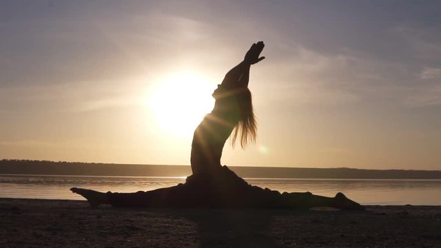 Silhouette of a beautiful yoga girl at sunrise on the beach. Hanumanasana, monkey pose. Sunrays lay on a body