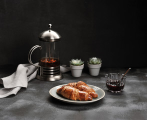 Two crispy croissants on a plate. Next a cup of jam and a teapot with tea. In the frame, cloth napkin. Dark background. Close-up.