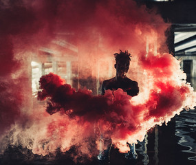 Young African man stands under bridge and holds colored red smoke bomb in his hands.