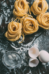 top view of raw tagliatelle pasta, sieve, egg and egg shell on table covered by flour