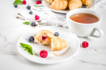 Sweet summer dessert, homemade baked mini croissants with berry jam, served with tea, fresh raspberries, blueberries and mint. On a white marble table, copy space