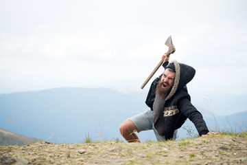 Man with beard on shouting face conquers top of mountain with axe, sky background. Survive in wild...