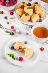 Sweet summer dessert, homemade baked mini croissants with berry jam, served with tea, fresh raspberries, blueberries and mint. On a white marble table, copy space