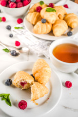 Sweet summer dessert, homemade baked mini croissants with berry jam, served with tea, fresh raspberries, blueberries and mint. On a white marble table, copy space