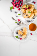 Sweet summer dessert, homemade baked mini croissants with berry jam, served with tea, fresh raspberries, blueberries and mint. On a white marble table, copy space top view