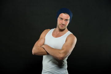 Man stand with strong arms folded. Macho in trendy vest and hat on dark background. Sportsman with bristle on serious face. Fashion style and trend. Sport and fitness