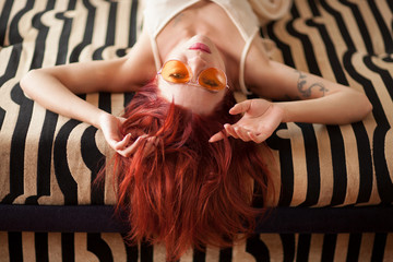 pretty young woman with red hair and sunglasses lies relaxed and laughing on the couch, emotion...