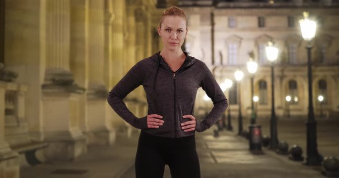 Confident Caucasian woman in gray hoodie doing power pose outside at night, Healthy determined young lady in exercise clothes posing proudly by old building at night, 4k
