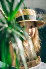 Smiling preschool girl in boater hat hiding behind the plant. Summer vacation concept. Vertical orientation