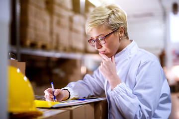 Young beautiful focused female worker is checking statistics papers on stack of boxes .