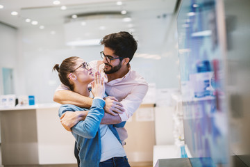 Young handsome joyful man is holding his girlfriend hugged from back as she is explaining him something.