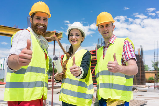 Portrait of three cheerful colleagues in a construction team looking at camera, while showing thumbs up as a gesture of agreement and confidence during work in progress