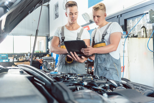 Experienced auto mechanic using a laptop for scanning and interpreting engine error codes next to an apprentice in a modern automobile repair shop