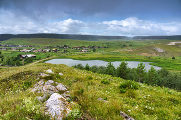 Obraz premium Heavy rain over the Ural village. Lake in the hills. Beautiful colorful sky. Russia