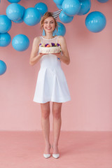 Young woman holding cake with macaroon decor on pink background with blue balls hanging. Full lenght portrait