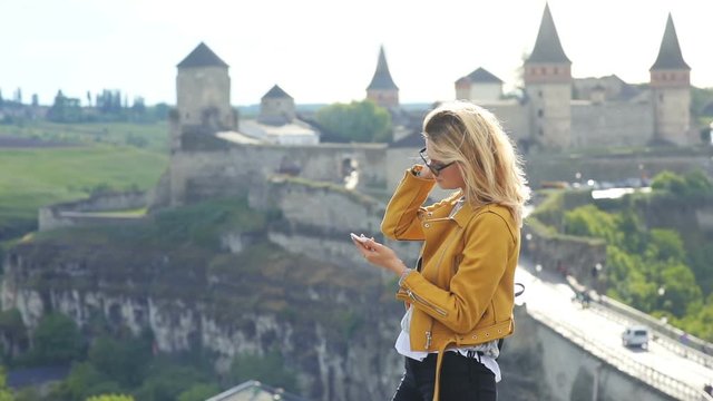 fashionable traveling girl with sunglasses in sunny spring day using smart phone cellphone outside with beautiful old brick castle on blur background culture tourism map app internet searching network