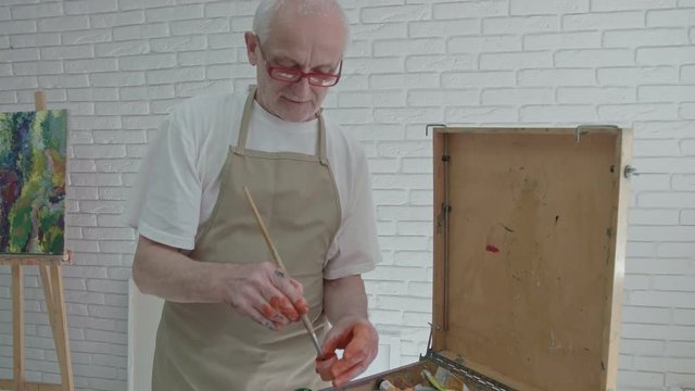 Old painter's hand taking paint with a brush, standing near big palette. Working in spacy atelier with white walls. Having paint spots on hands. Feeling inspired, loving work.