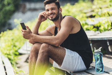 smiling sportsman with bottle of water listening music with smartphone and earphones on bench at sport playground