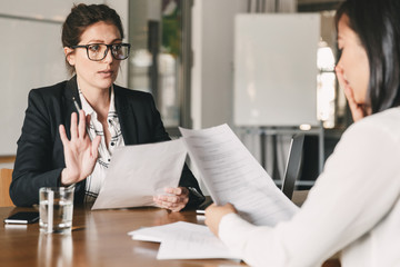Image of nervous unsure asian woman talking and negotiating with businesswoman, while sitting at...