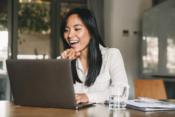 Photo of cheerful asian woman 20s wearing white shirt laughing and pointing finger at screen of laptop, while speaking or chatting on video call in office