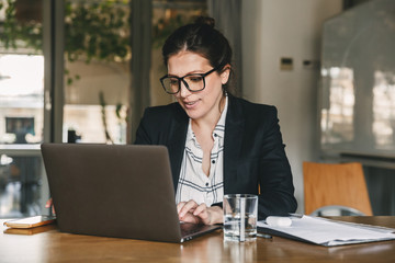 Photo of caucasian boss woman 30s wearing formal clothing and eyeglasses smiling while sitting at...