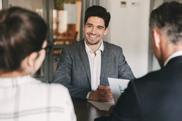 Business, career and placement concept - young caucasian man smiling, while sitting in front of directors during corporate meeting or job interview - obrazy, fototapety, plakaty