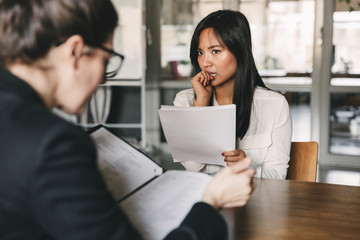 Image of concentrated tense asian woman looking at businesswoman, while sitting at table in office during job interview - business, career and recruitment concept - obrazy, fototapety, plakaty