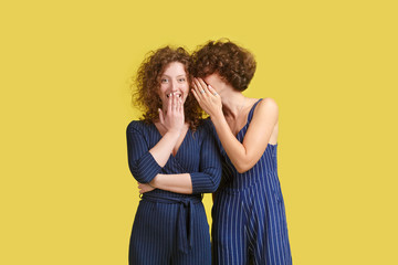 Handsome young curly European female in blue overall whispering something into ear of stylish redhead girl, sharing latest gossip. Ginger Woman looking in shock and surprise with mouth wide open