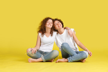 Two charming curly sisters or friends in white t-shirt and trendy jeans are sitting on studio yellow floor, hugging each other, spending time together fun. Family Relationships and Values
