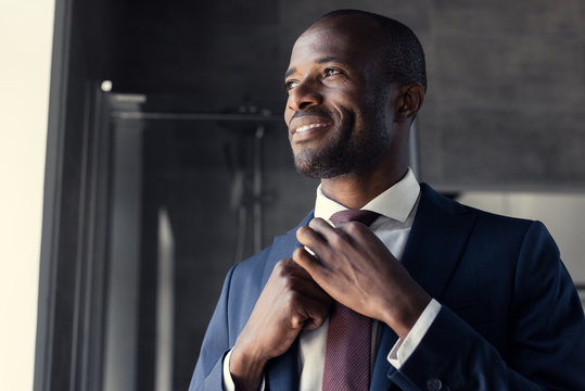 smiling young businessman putting on his tie at bathroom and looking away
