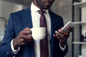 cropped shot of handsome young businessman with cup of coffee using smartphone