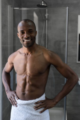 Fototapeta na wymiar handsome bare-chested african american man in towel standing with hands on waist and smiling at camera in bathroom