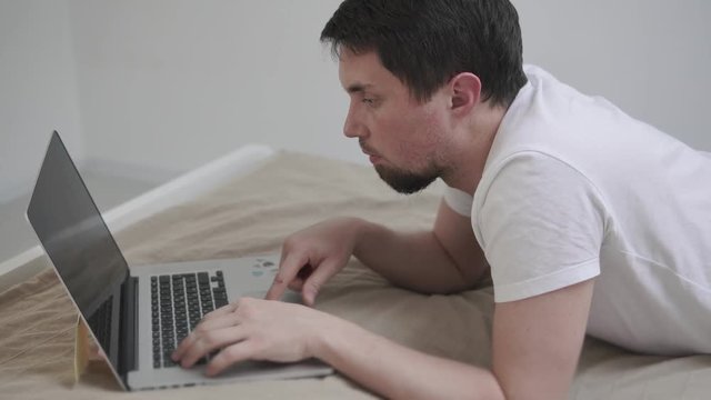 Close up shot of a man lying in bed with a laptop. Man is surfing the web and sitting on social networks. Scrolling the feed and smiling.