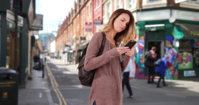 Young woman walks through hipster neighborhood using smartphone for directions, Pretty Caucasian girl wandering down urban street using mobile phone, 4k