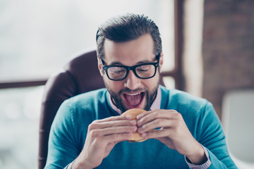 Close up portrait of cheerful busy excited handsome hungry manager doing to bite yummy appetizing cheeseburger in his hands sitting on armchair at workstation