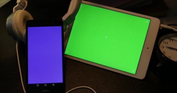 Dolly shoot of blue screen cellphone and green screen tablet computer with headphone on desk at night 