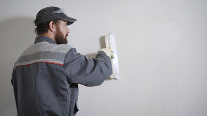 plasterer is wearing work clothes and cap is aligning wall, using white decorative putty, repair in...