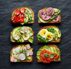 Avocado sandwiches, toasts with various vegetarian toppings on a black stone background, top view
