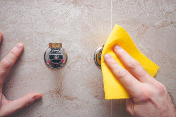 The man is rubbing the tile and the water taps on the wall in the bathroom