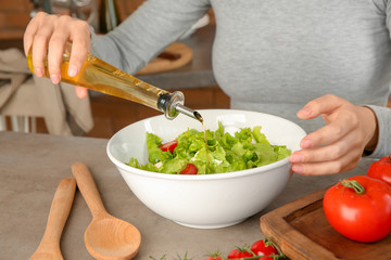 Woman adding olive oil into bowl with fresh vegetable salad on table