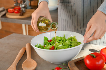 Woman adding olive oil into bowl with fresh vegetable salad on table